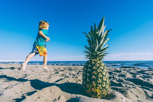 enfant courant sur la plage avec au premier plan un ananas posé dans le sable